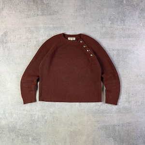 May include: A rust-colored sweater with a crew neck and long sleeves. The sweater features a row of four decorative buttons on the shoulder. The knitwear is a solid color and has a ribbed texture.