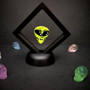 Op de afbeelding: Een felgeel gitaarplectrum met een zwarte alien-gezichtsgrafiek, zwevend in een zwart ruitvormig displayframe. Zes kleine, doorschijnende, veelkleurige schedelvormige beeldjes zijn in een halve cirkel rond de basis gerangschikt.