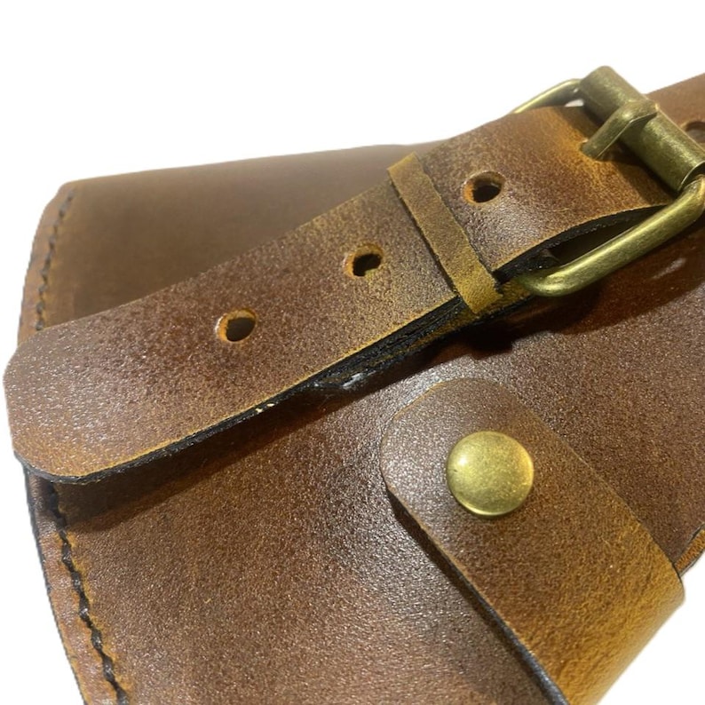 May include: Close-up of a brown leather wallet with a buckle closure. The wallet features a distressed leather texture and a brass-colored buckle and stud. The design suggests a rustic or vintage aesthetic.