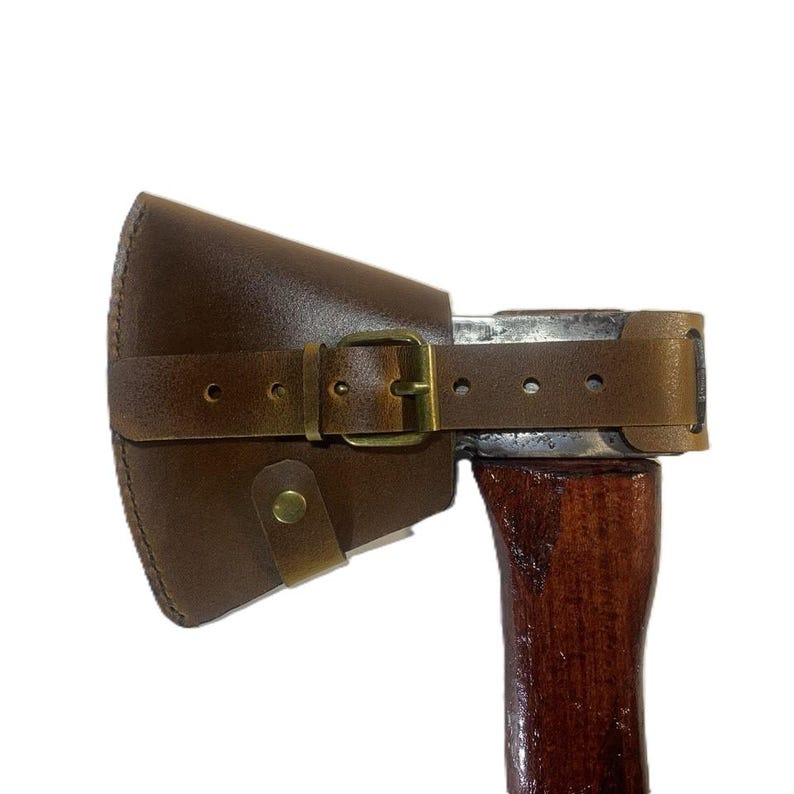 May include: A brown leather axe sheath with a buckle closure. The sheath covers the axe head and is secured with a leather strap and a brass buckle. The axe handle is made of dark wood.