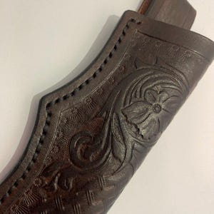 May include: Close-up of a brown leather sheath with detailed floral and geometric embossed patterns. The sheath has a stitched border and a belt loop. The leather is a rich, dark brown colour.