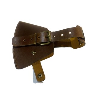 May include: Brown leather axe sheath with a buckle closure and a strap. The sheath has a riveted design and a small flap with a snap closure. The leather has a worn appearance.