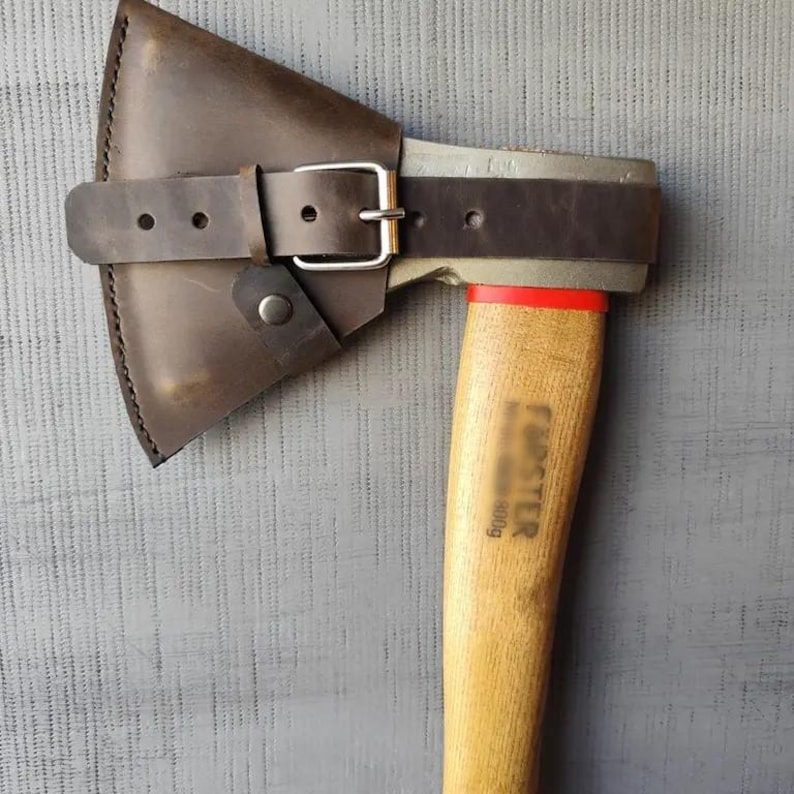 May include: Axe with a wooden handle and a metal head, featuring a brown leather sheath secured by a belt with a silver buckle. The handle has the word "FORSTER" printed on it.