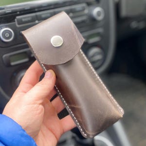 Puede incluir: Una funda de cuero marrón con cierre de solapa asegurado con un broche plateado. La funda tiene costuras blancas a lo largo de los bordes y parece hecha a mano. La funda se sostiene en una mano.