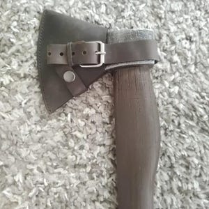 May include: Axe with a dark brown wooden handle and a silver-colored head. The axe head is covered with a dark brown leather sheath secured by a matching leather strap and a silver buckle. The axe is designed for chopping wood.