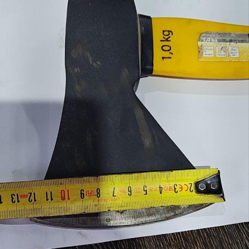 May include: A black axe head with a yellow handle. The handle is marked with "1,0 kg". A yellow tape measure shows the axe blade's length. The axe is a tool for chopping wood or other materials.