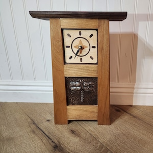 May include: A handcrafted wooden side table with a square clock face and a dragonfly design. The clock face is light-colored wood with a compass-like design. The table is made of dark and light brown wood.