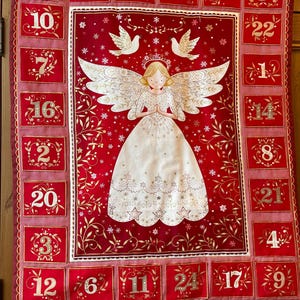May include: Red and gold fabric advent calendar featuring an angel design. The angel has large wings and a white dress. The calendar has 24 numbered pockets, each with a gold number, perfect for the festive season.