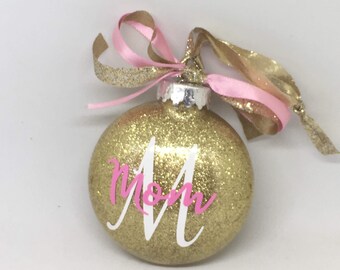 May include: Gold glitter Christmas ornament with a pink ribbon and the word "Mom" in white lettering.