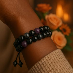 May include: Two beaded bracelets, one black and one with purple beads, are worn on a wrist. The bracelets are made of round beads and are strung on a black cord. The background is blurred, with a hint of flowers.