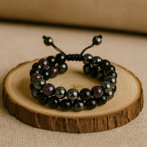 May include: Two-strand beaded bracelet featuring black and purple beads. The bracelet is displayed on a small, round wooden slice, set against a neutral background. The beads have a polished finish.