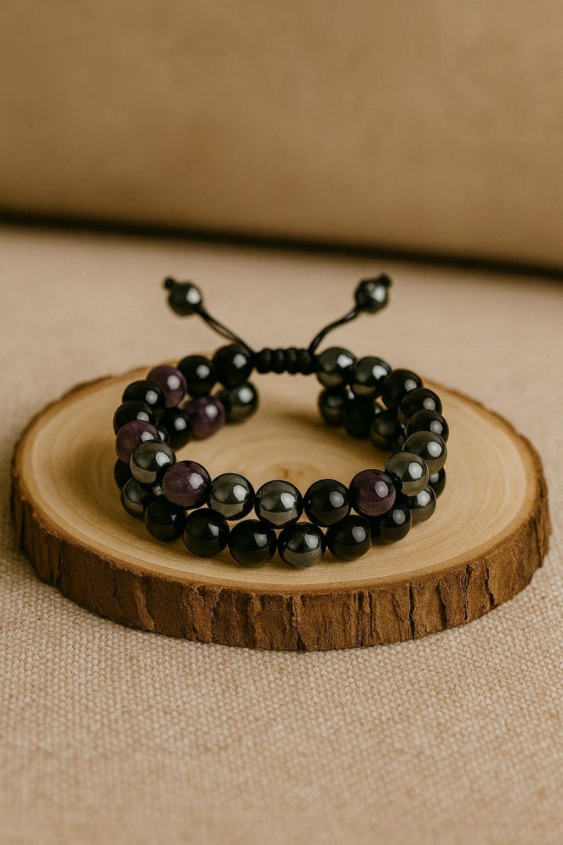 May include: Two-strand beaded bracelet featuring black and purple beads. The bracelet is displayed on a small, round wooden slice, set against a neutral background. The beads have a polished finish.