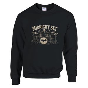 May include: A black long-sleeve sweatshirt featuring a graphic print of a skeleton playing a drum set. The text MIDNIGHT SET is printed in a distressed beige font above the skeleton. The bass drum features a circular bat logo in the center.