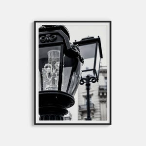 Può includere: Stampa incorniciata in bianco e nero con un primo piano di un lampione d'epoca. La lampada presenta dettagli decorativi e un involucro di vetro. Una seconda lampada è visibile sullo sfondo. L'immagine cattura un'estetica architettonica classica.