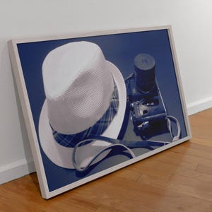 Op de afbeelding: Een ingelijste print met een witte fedora hoed met een geruite binnenkant, een vintage zwarte camera en een zonnebril. De objecten zijn geplaatst tegen een donkerblauwe achtergrond, wat een stilleven creëert.