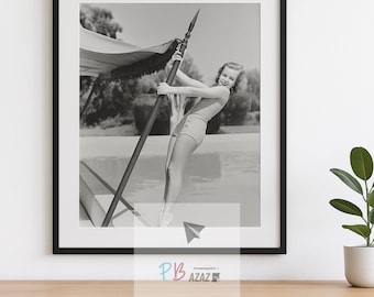 May include: Black and white framed print of a vintage photograph. The image features a woman in a swimsuit holding a pole with a canopy, posing near a pool. The print is displayed on a wooden surface.