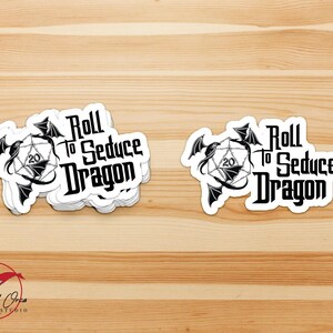 May include: White stickers with the text "Roll to Seduce Dragon" in a gothic font, featuring a dragon and a twenty-sided die. The stickers are on a light brown wooden surface. The Red Orca Studio logo is in the bottom left corner.