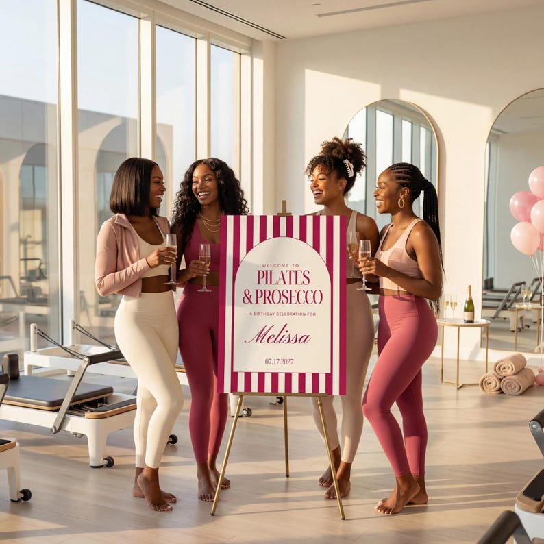 May include: Four women in workout attire holding champagne flutes, gathered around a sign that reads "Pilates & Prosecco" with the name "Melissa" and the date 07.17.2022. The sign is on a gold easel. The setting is a bright, modern studio.