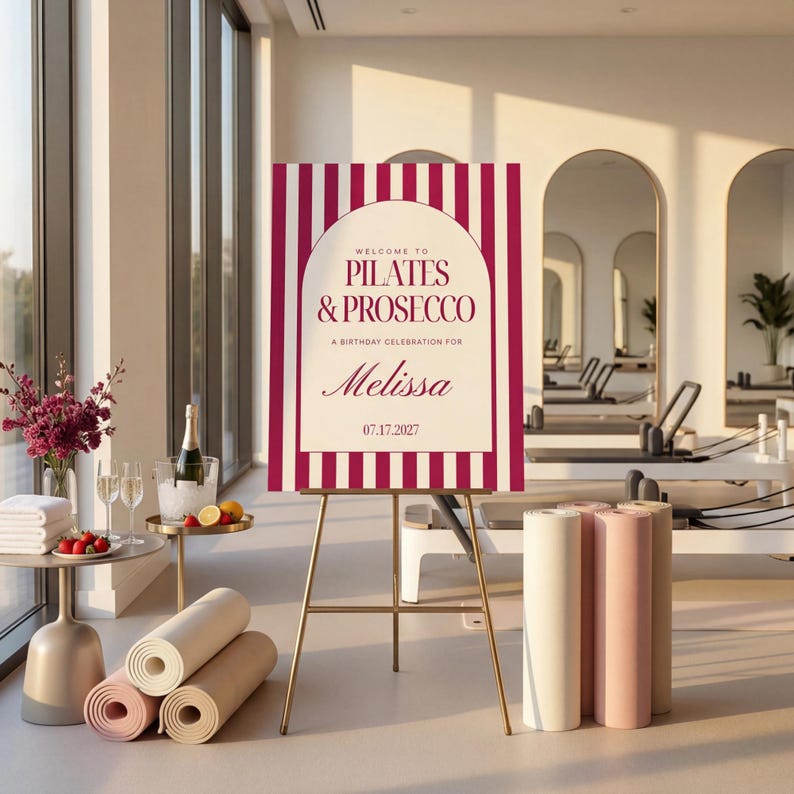 May include: A gold easel displays a sign with burgundy and white stripes. The sign reads "Welcome to Pilates & Prosecco, A Birthday Celebration for Melissa, 07.17.2027." Rolled yoga mats in beige and pink are in the foreground.