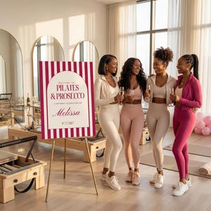 May include: A sign with the text "WELCOME TO PILATES & PROSECCO" and the name "Melissa" is displayed on a gold easel. Four women in athletic wear are holding champagne flutes. The sign has a burgundy and white striped design.