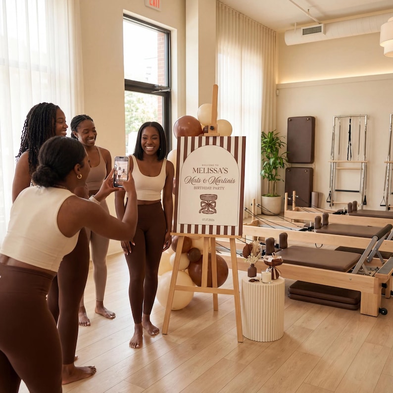 May include: A group of women pose for a photo in a pilates studio. A sign on an easel reads "Melissa's Mats & Martinis Birthday Party." The women are wearing neutral-toned athletic wear. Balloons and pilates equipment are visible in the background.