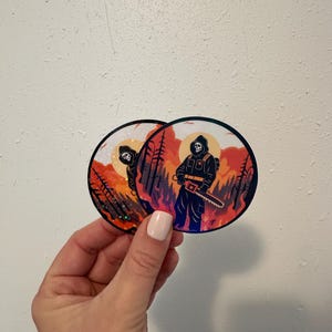 Può includere: Due adesivi rotondi raffiguranti una figura nera con cappuccio che tiene una motosega, sullo sfondo di un tramonto infuocato con alberi. Gli adesivi hanno una finitura olografica iridescente.
