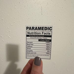 Puede incluir: Una pegatina blanca con texto negro que dice "PARAMEDIC Nutrition Facts". La pegatina enumera los valores diarios de sarcasmo, dolor de espalda, cafe&iacute;na, sirenas, empat&iacute;a y "Saves your ass". La pegatina est&aacute; sujeta por una mano con esmalte de u&ntilde;as gris.