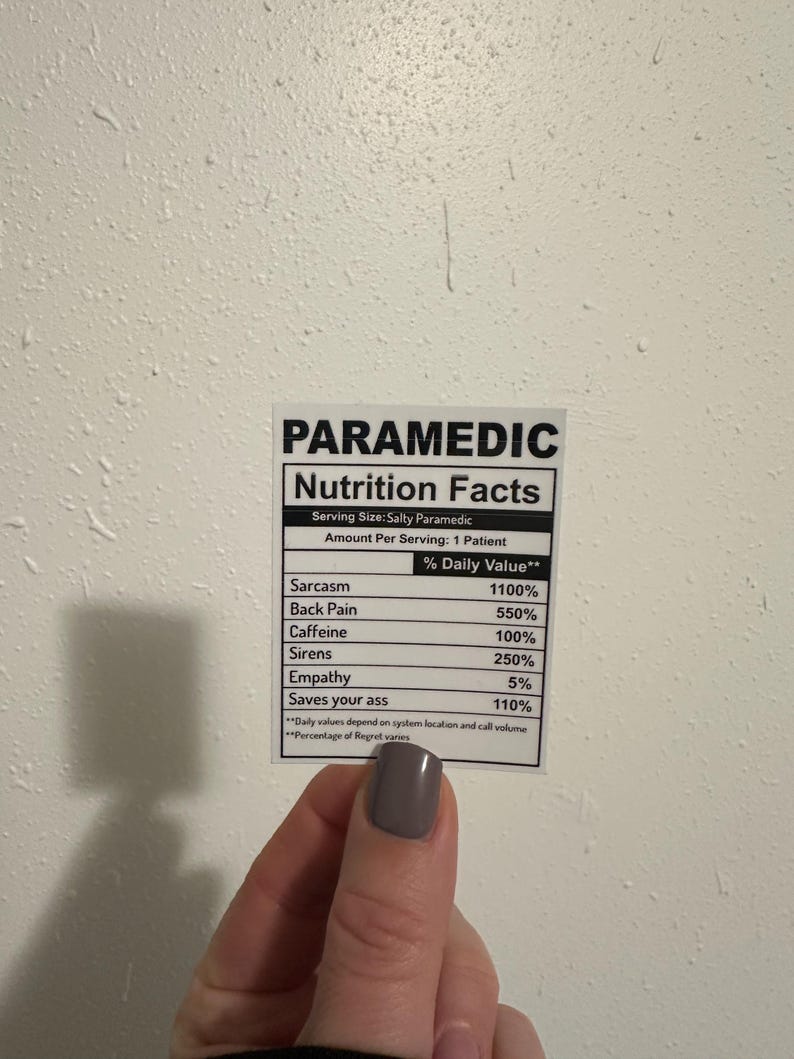Puede incluir: Una pegatina blanca con texto negro que dice "PARAMEDIC Nutrition Facts". La pegatina enumera los valores diarios de sarcasmo, dolor de espalda, cafe&iacute;na, sirenas, empat&iacute;a y "Saves your ass". La pegatina est&aacute; sujeta por una mano con esmalte de u&ntilde;as gris.