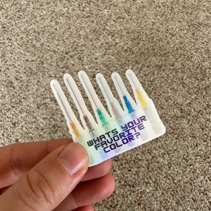 May include: A sticker with a holographic finish featuring six medical needles with colored tips. The sticker has the text "WHAT'S YOUR FAVORITE COLOR?" in a blue and purple gradient. The needles have orange, green, pink, blue, and yellow tips.
