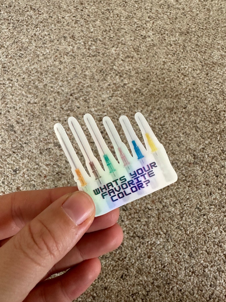 May include: A sticker with a holographic finish featuring six medical needles with colored tips. The sticker has the text "WHAT'S YOUR FAVORITE COLOR?" in a blue and purple gradient. The needles have orange, green, pink, blue, and yellow tips.