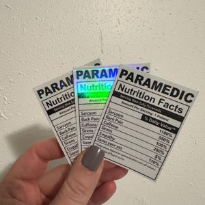 Puede incluir: Tres pegatinas con fondo blanco y texto negro que dice "PARAMEDIC Nutrition Facts". Las pegatinas enumeran datos humor&iacute;sticos de "% Daily Value" como "Sarcasm" y "Caffeine". Las pegatinas se sostienen en una mano.