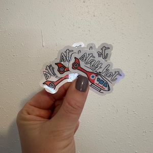 May include: Holographic stickers featuring a red and white helicopter design with the text "stay hot." The stickers have a reflective, iridescent finish. The helicopter design includes a red tail and a blue and white body.
