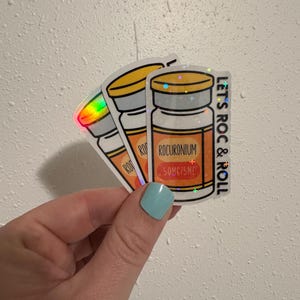Może przedstawiać: Trzy holograficzne naklejki przedstawiające rysunkowy flakon leku. Naklejki mają żółtą nakrętkę i pomarańczową etykietę z napisem "ROCURONIUM 50MG/5ML" i "LET'S ROC & ROLL".