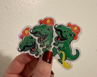 Puede incluir: Tres pegatinas de dinosaurios verdes con bigotes negros y sombreros de bombero rojos. Cada dinosaurio tiene la boca abierta y una manguera amarilla. Las pegatinas se sostienen en una mano sobre un fondo blanco.