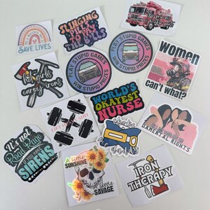 May include: A collection of various stickers with different designs and text. The stickers feature phrases like "Save Lives," "Play Stupid Games," and "World's Okayest Nurse." Other designs include a skull with sunflowers, a fire truck, and a dumbbell.