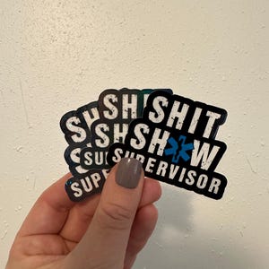 Könnte beinhalten: Mehrere schwarz-weiße Aufkleber mit dem Text "SHIT SUPERVISOR" in fetter Schrift. Die Aufkleber zeigen auch ein blaues Medizinsymbol. Die Aufkleber werden in einer Hand mit neutralfarbenem Nagellack gehalten.