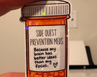 Può includere: Un adesivo fustellato a forma di flacone di medicinali arancione con tappo bianco. L'etichetta riporta il testo "SIDE QUEST PREVENTION MEDS" seguito da "Because my brain has better ideas than my focus." e un piccolo simbolo a cuore.