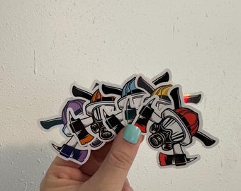Puede incluir: Una colección de pegatinas de cascos de bombero coloridos con hachas. Las pegatinas presentan cascos en varios colores, incluyendo rojo, azul, amarillo y morado, con detalles en blanco y negro. Las pegatinas se sostienen en una mano sobre un fondo blanco.