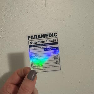 Puede incluir: Una pegatina con una etiqueta "PARAMEDIC Nutrition Facts". La etiqueta enumera los valores diarios de sarcasmo, dolor de espalda, cafe&iacute;na, sirenas, empat&iacute;a y Saves your ass. La pegatina est&aacute; sujeta por una mano.