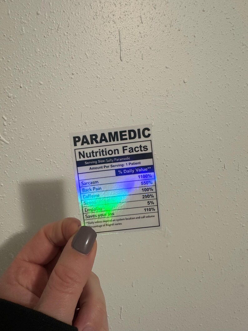Puede incluir: Una pegatina con una etiqueta "PARAMEDIC Nutrition Facts". La etiqueta enumera los valores diarios de sarcasmo, dolor de espalda, cafe&iacute;na, sirenas, empat&iacute;a y Saves your ass. La pegatina est&aacute; sujeta por una mano.