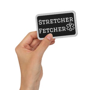 May include: A rectangular black patch with a white embroidered border. The text STRETCHER FETCHER is embroidered in white capital letters, accompanied by a white Star of Life medical symbol on the right side of the patch against the black background.
