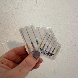 May include: Clear stickers with a syringe design and the text "WHAT'S YOUR FAVORITE COLOR?" The syringes have colorful tips. The stickers are held in a hand against a textured white background.
