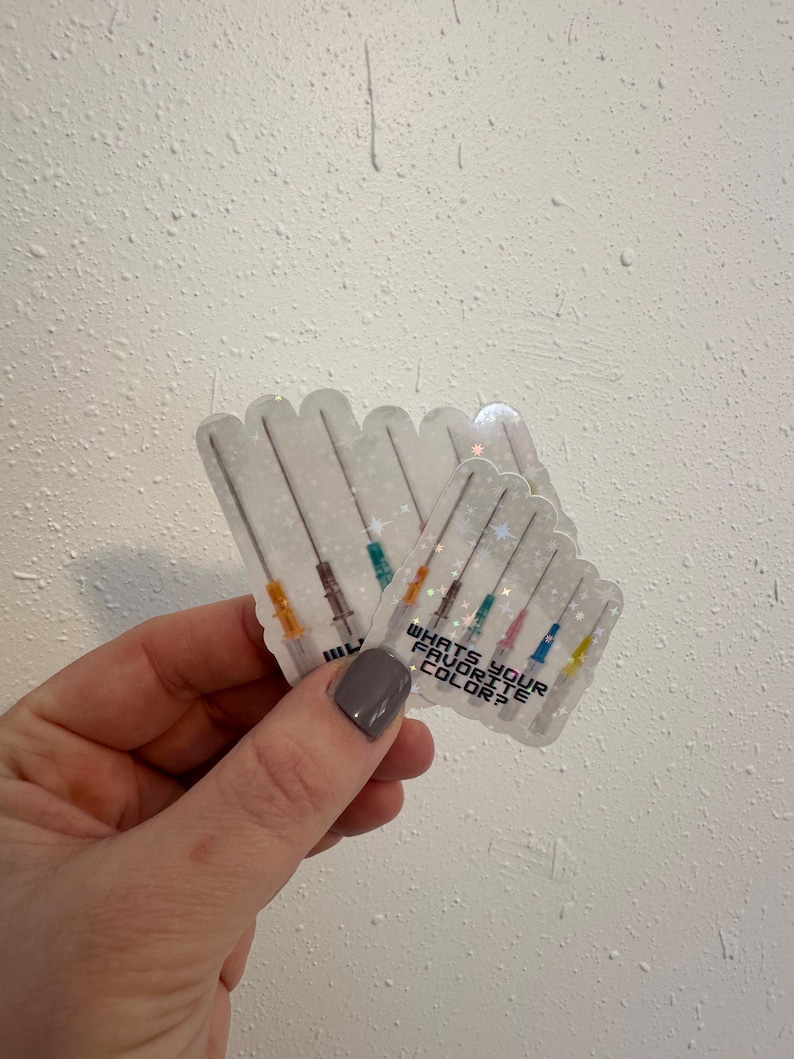 May include: Clear stickers with a syringe design and the text "WHAT'S YOUR FAVORITE COLOR?" The syringes have colorful tips. The stickers are held in a hand against a textured white background.