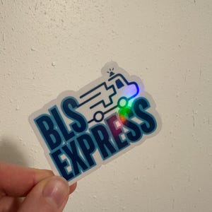 May include: A sticker with the words "BLS EXPRESS" in a bold, blue font. The sticker features a line drawing of an ambulance with a flashing light. The sticker has a holographic, iridescent effect.