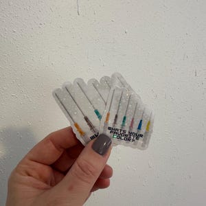 May include: Clear stickers featuring syringe illustrations with colorful needles. The stickers have the text "WHAT'S YOUR FAVORITE COLOR?" printed on them. The stickers are held in a hand against a white textured background.
