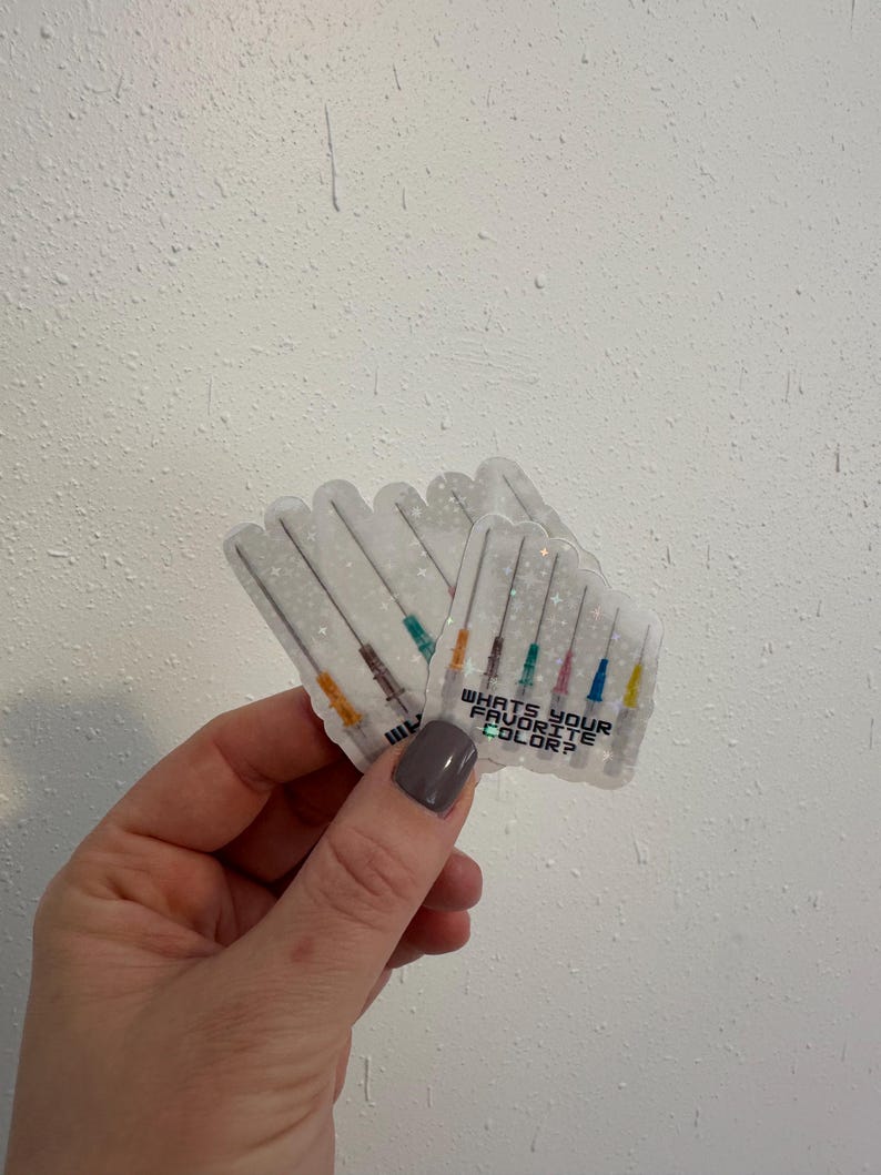 May include: Clear stickers featuring syringe illustrations with colorful needles. The stickers have the text "WHAT'S YOUR FAVORITE COLOR?" printed on them. The stickers are held in a hand against a white textured background.