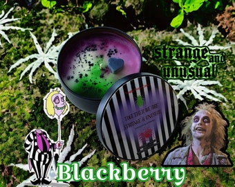 Può includere: Un barattolo di candela rotondo nero con un coperchio con un design a strisce bianche e nere. La cera della candela è un mix di viola e verde con un accento a forma di cuore. Il testo "Together We Are Strange & Unusual" è visibile.