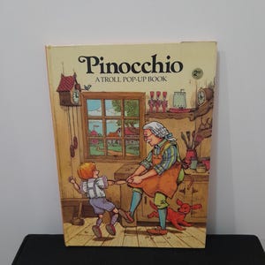 May include: A vintage copy of "Pinocchio: A Troll Pop-Up Book" with a colorful illustration of a scene from the story. The cover features the title and a scene with a woman and a boy. The book is set against a neutral background.