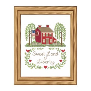 May include: Cross-stitch artwork in a wooden frame. The image features a red house with an American flag, trees, and the text "Sweet Land of Liberty" surrounded by a floral wreath. The frame is a light brown color.