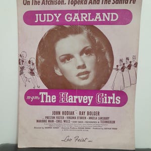 May include: Vintage sheet music cover for "On The Atchison, Topeka And The Santa Fe" featuring Judy Garland. The cover has a pink and white color scheme with text and a portrait of Garland. The title "The Harvey Girls" is also displayed.
